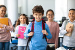 Return Back To School. Smiling small boy wearing backpack posing in classroom, diverse group of happy international classmates standing in line in the blurred background, studying and learning